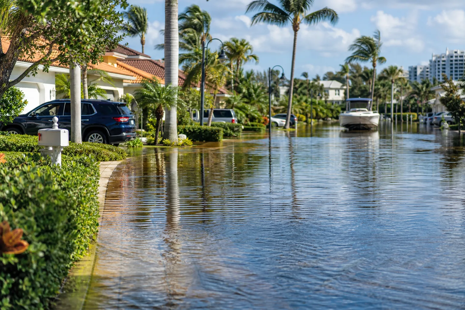 Flooded residential street in Florida with palm trees, luxury homes, parked SUVs, and a boat floating on floodwaters